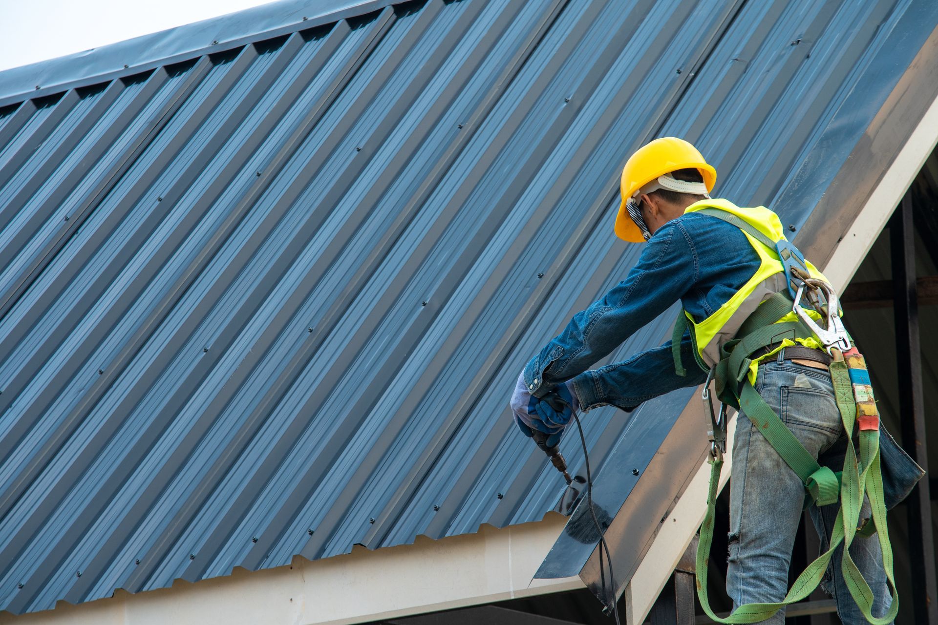 Worker in yellow hard hat repairs a blue metal roof with power tools.