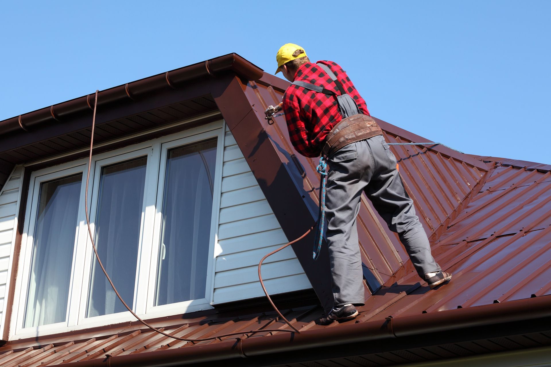 Worker on a red roof cleaning gutters with a hose, wearing a yellow hard hat and safety gear.