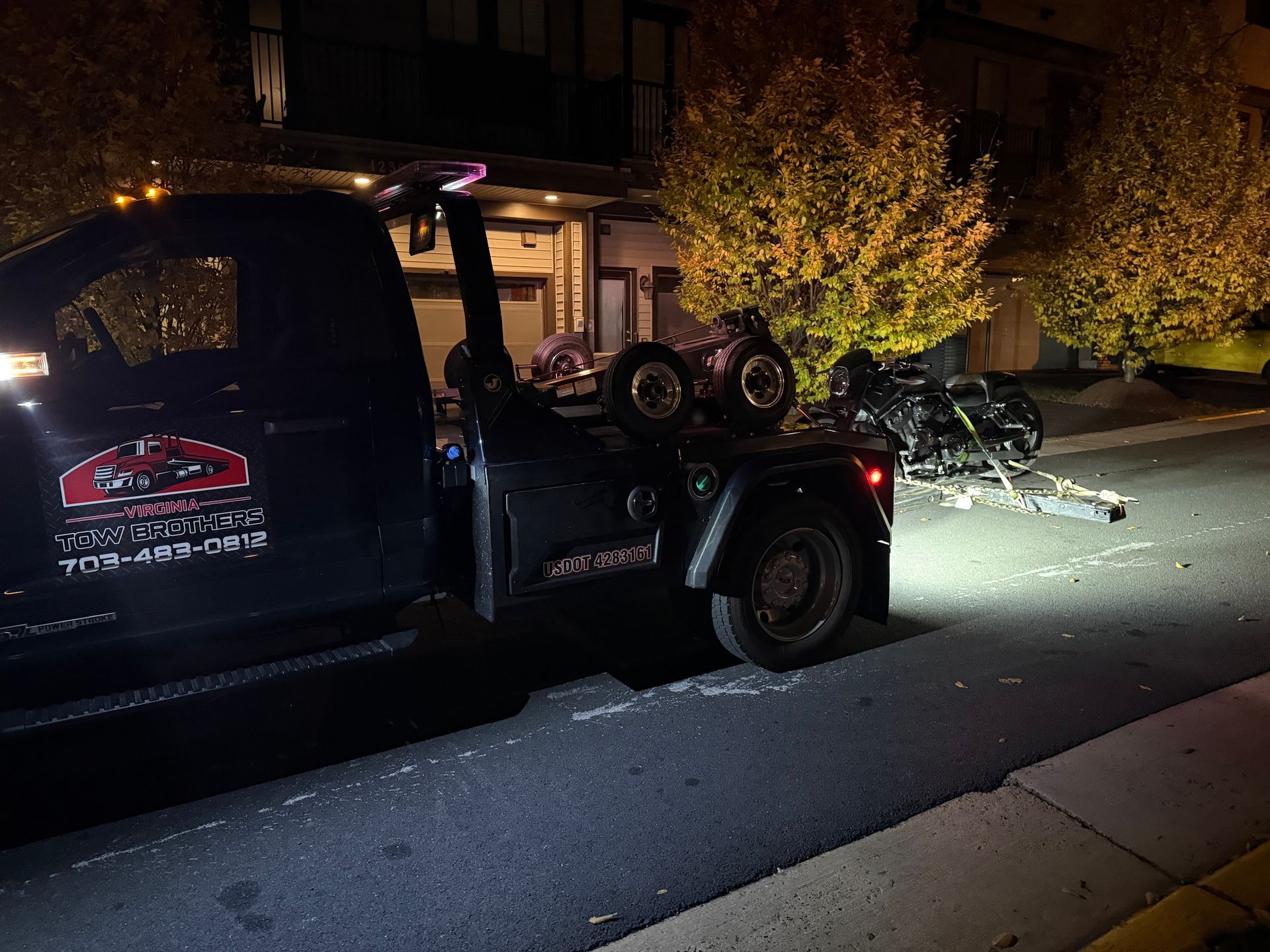 A tow truck is towing a motorcycle down a street at night.