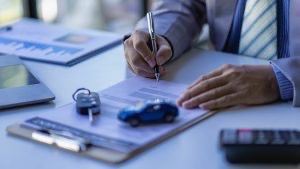 A man in a suit and tie is writing on a clipboard next to a toy car.