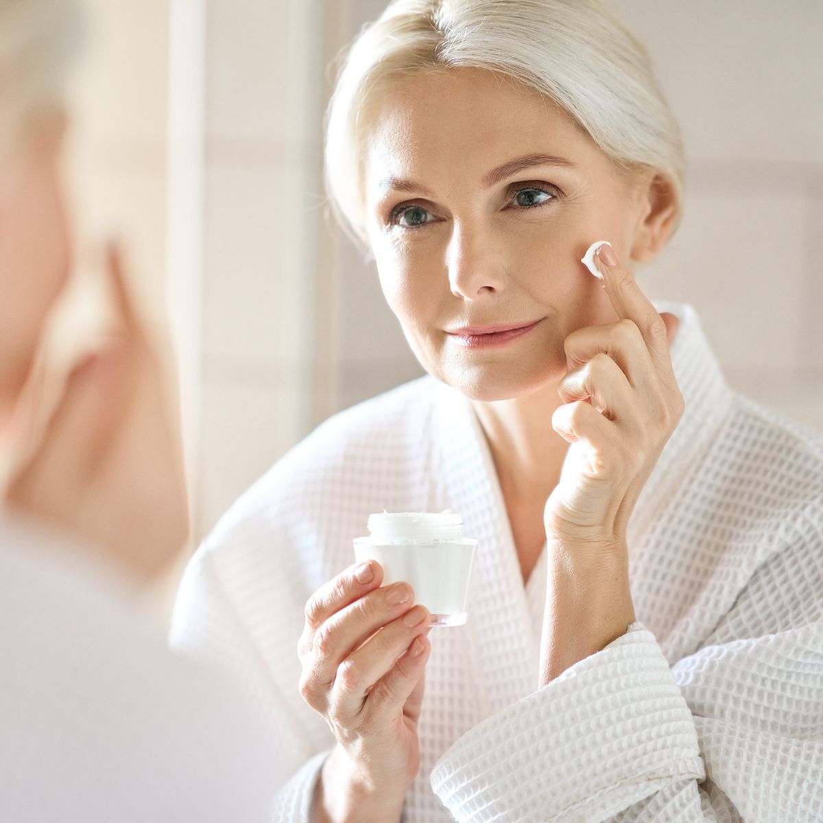 Woman in a white robe applying cream to her face in front of a mirror.