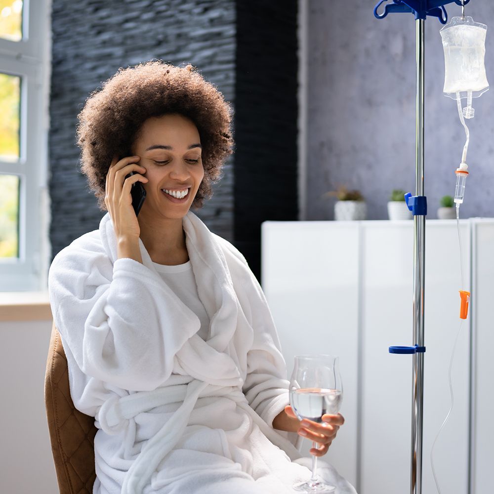 Woman in a robe smiling while on the phone, receiving IV drip therapy, holding water glass.