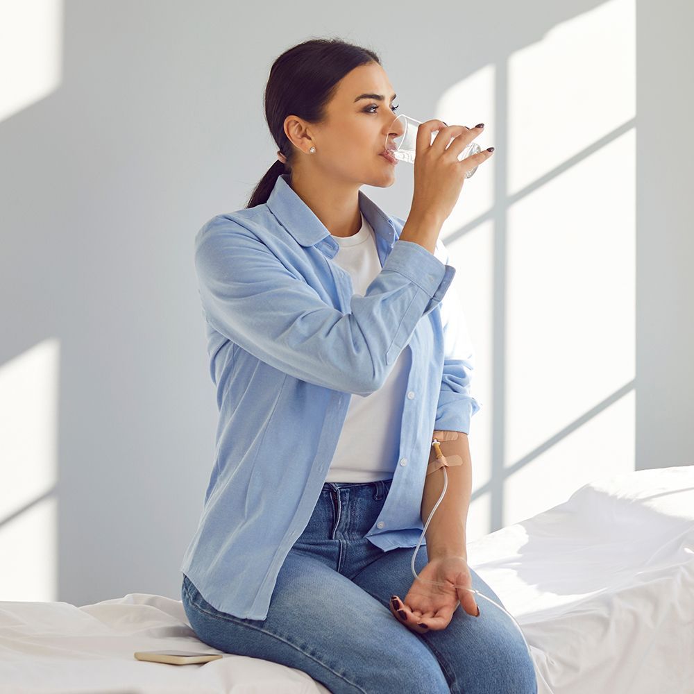 Woman drinks water while receiving IV fluids, seated on a bed. Bright room, blue shirt, jeans.