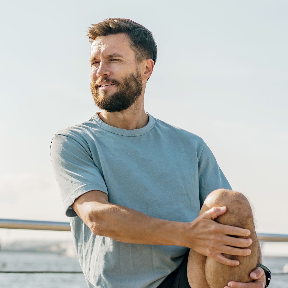 Man stretches leg outdoors near water, looking away.