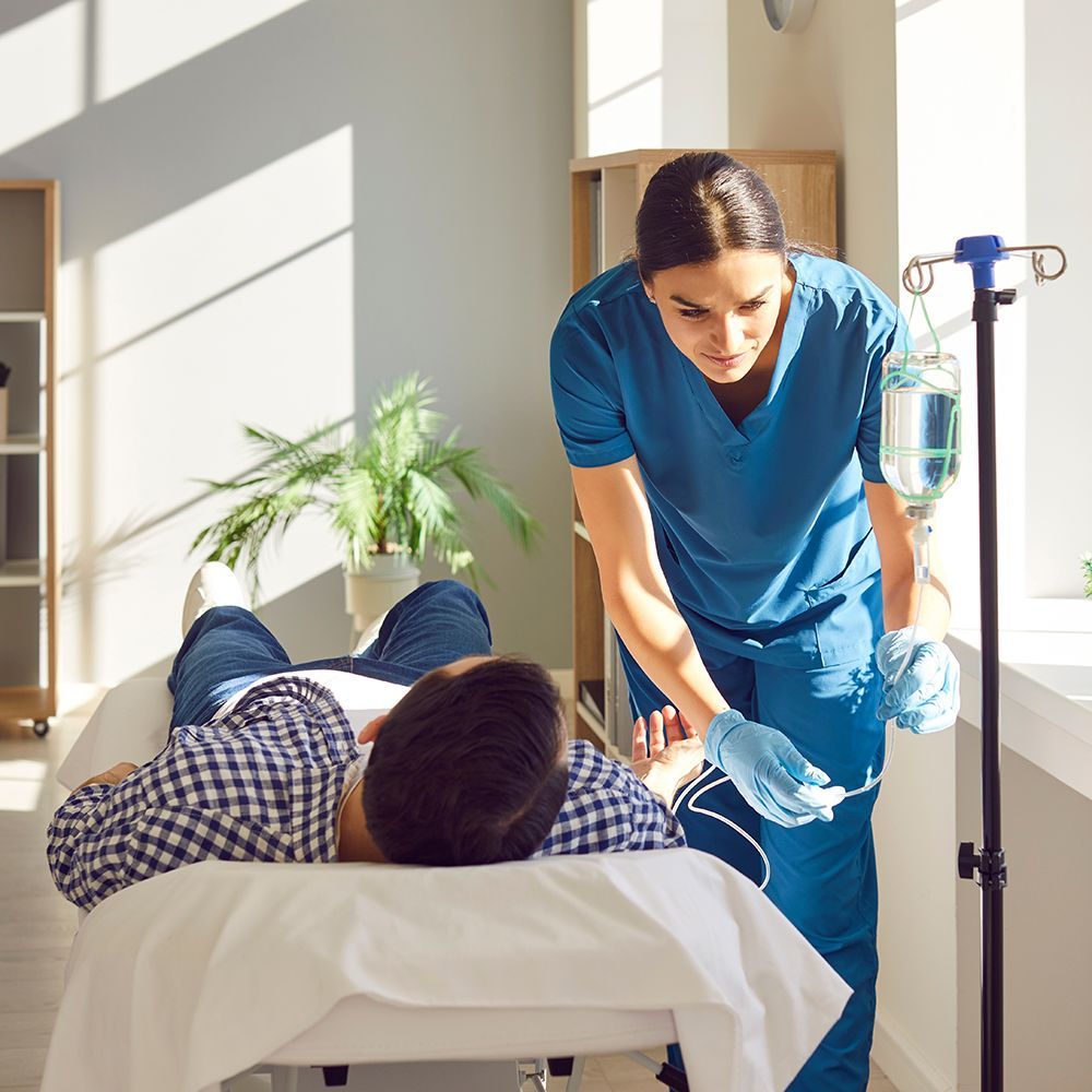 Nurse adjusts IV drip for a patient lying on a bed in a sunny room.