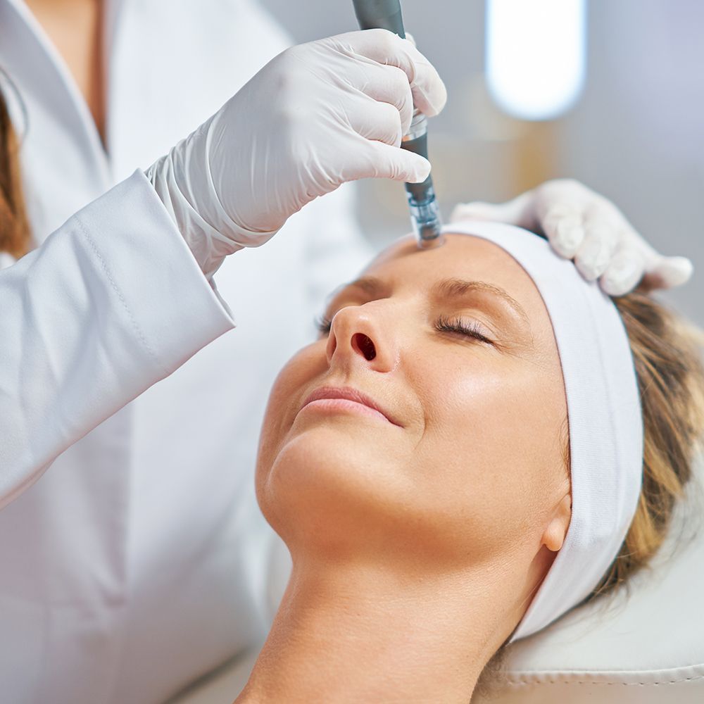 Person receiving microneedling treatment on forehead at a clinic. Blue gloves, closed eyes.
