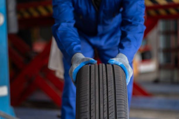 A man in blue overalls is holding a tire in a garage.