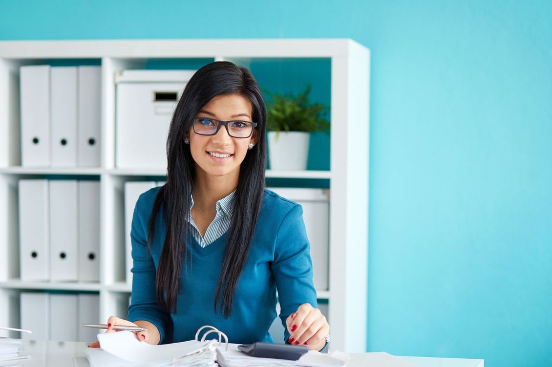 A woman is sitting at a desk in an office using a calculator.