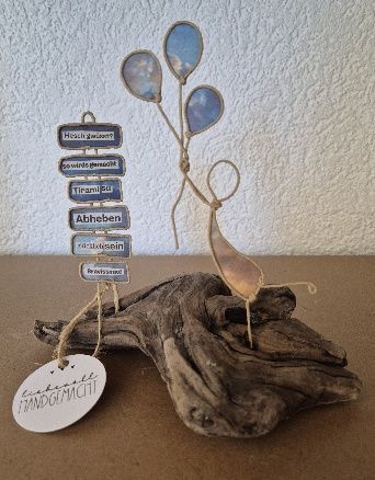 Buntglasbild einer Person mit Luftballons, befestigt an Treibholz, mit übereinander gestapelten Worttafeln.