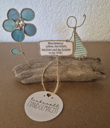 Blaue Blume und Drahtfigur auf Treibholz mit einem Schild und einem Anhänger mit der Aufschrift „Handgemacht“.