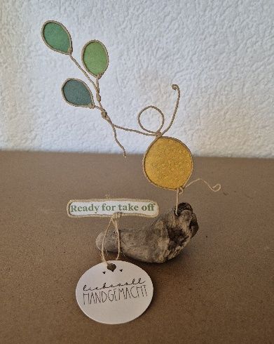 Drahtfigur mit Luftballons auf einem Steinsockel; Aufschrift: „Bereit zum Abheben“ und „Handgefertigt“.