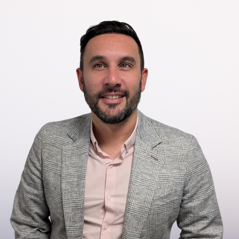 A man with a beard wearing a light pink shirt and gray blazer smiles at the camera against a white background.