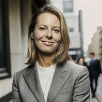 Woman in a grey blazer smiles at the camera; blurred background of a city street and people.