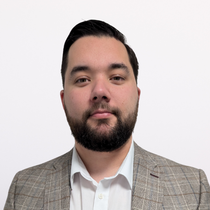 Man with dark hair and beard wearing a plaid blazer and white shirt, facing the camera against a white background.