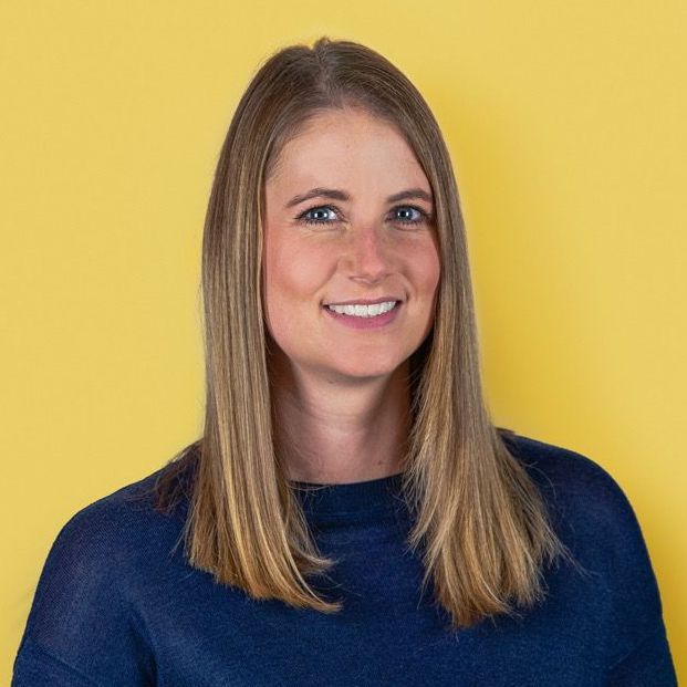Woman with shoulder-length blonde hair smiling, wearing a navy blue sweater against a yellow background.