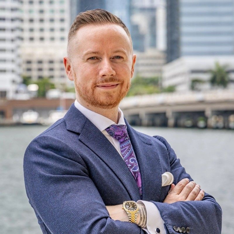 Man in blue suit with paisley tie, standing by water in a city. He is smiling with arms crossed.