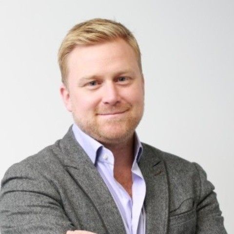 A smiling man with blond hair and a beard wearing a gray blazer over a light purple shirt.