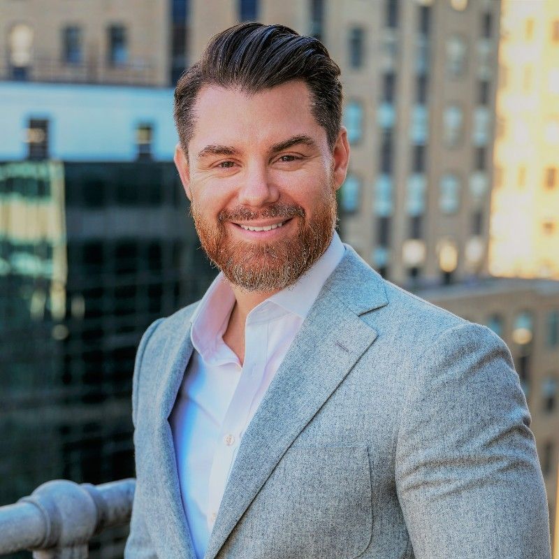A smiling man with a beard in a grey blazer, standing outside with a city backdrop.