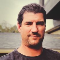 A man with short, dark hair and a beard looks towards the viewer with a neutral expression, outside a house.