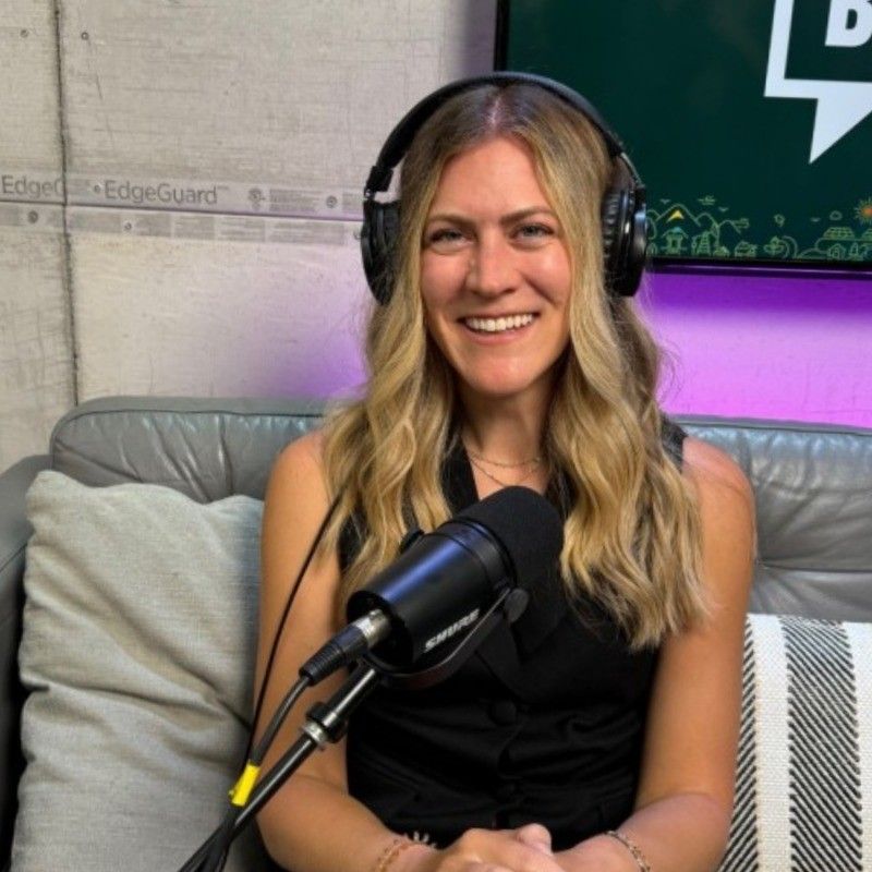 Blond woman with headphones smiles into a microphone, seated on a gray couch in a podcast studio.