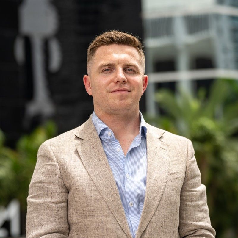 A man with short brown hair wearing a light brown blazer and light blue shirt smiles outdoors near a modern building.