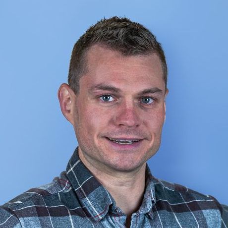 A man with short brown hair wearing a plaid shirt smiles at the camera against a light blue background.