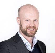 A smiling, bald man with a reddish-brown beard in a blazer and collared shirt against a white background.