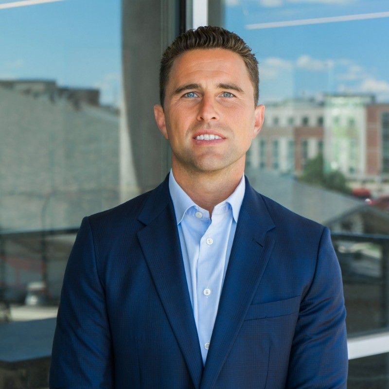 Man in a blue suit and open-collared shirt smiling, standing in front of a window with a city view.