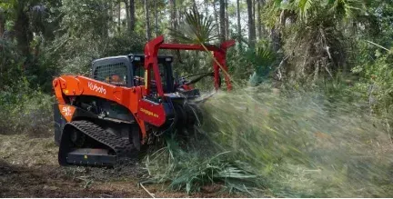 Orange Kubota skid steer mulching vegetation in a wooded area, spraying debris.