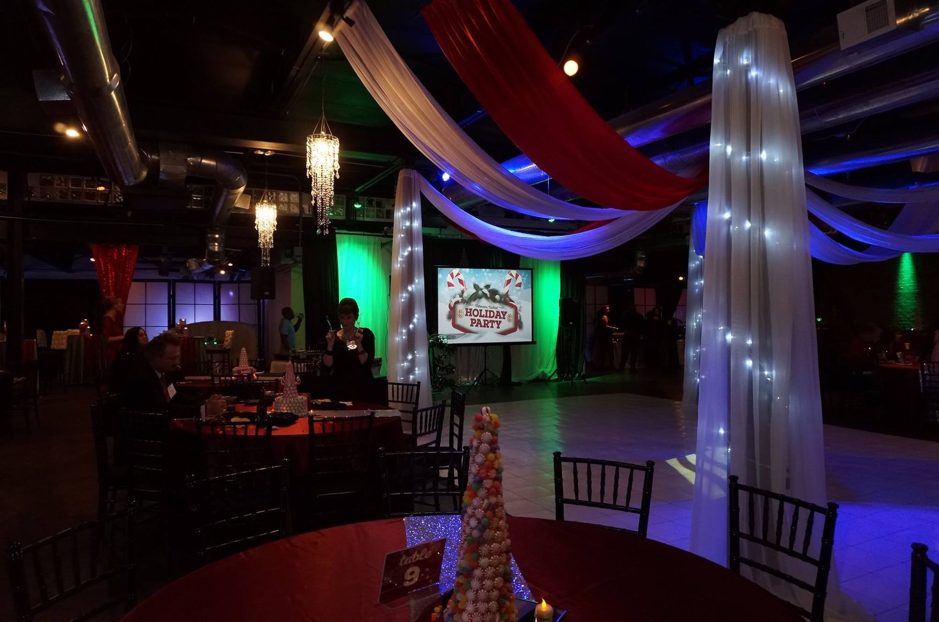 A large room with tables and chairs and a large screen hanging from the ceiling.
