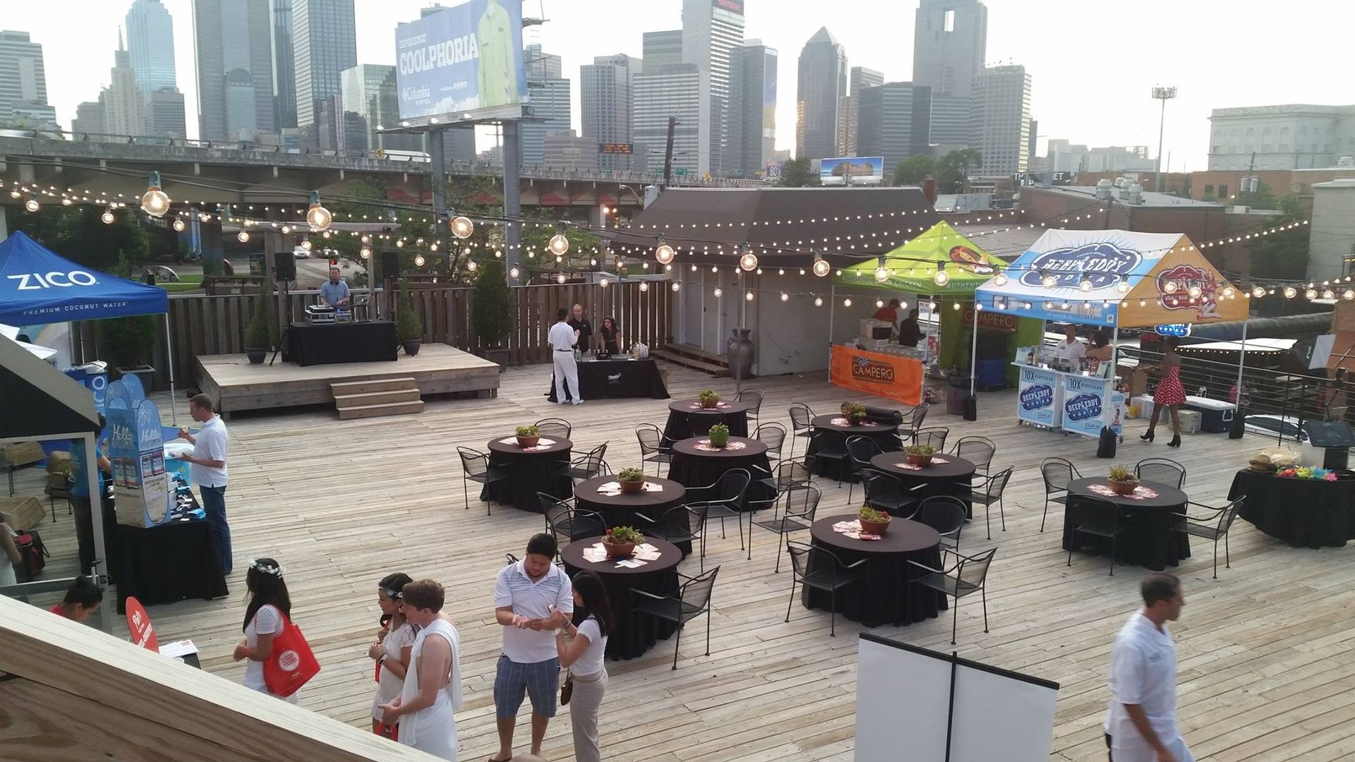 A group of people are standing around tables and chairs in front of a city skyline.