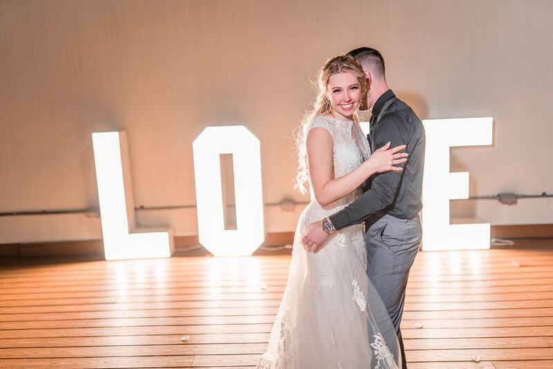 A bride and groom are dancing in front of a large love sign.