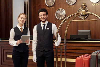 Two hotel staff smiling at the camera, one holding a tablet, next to a luggage cart and reception desk with clocks.