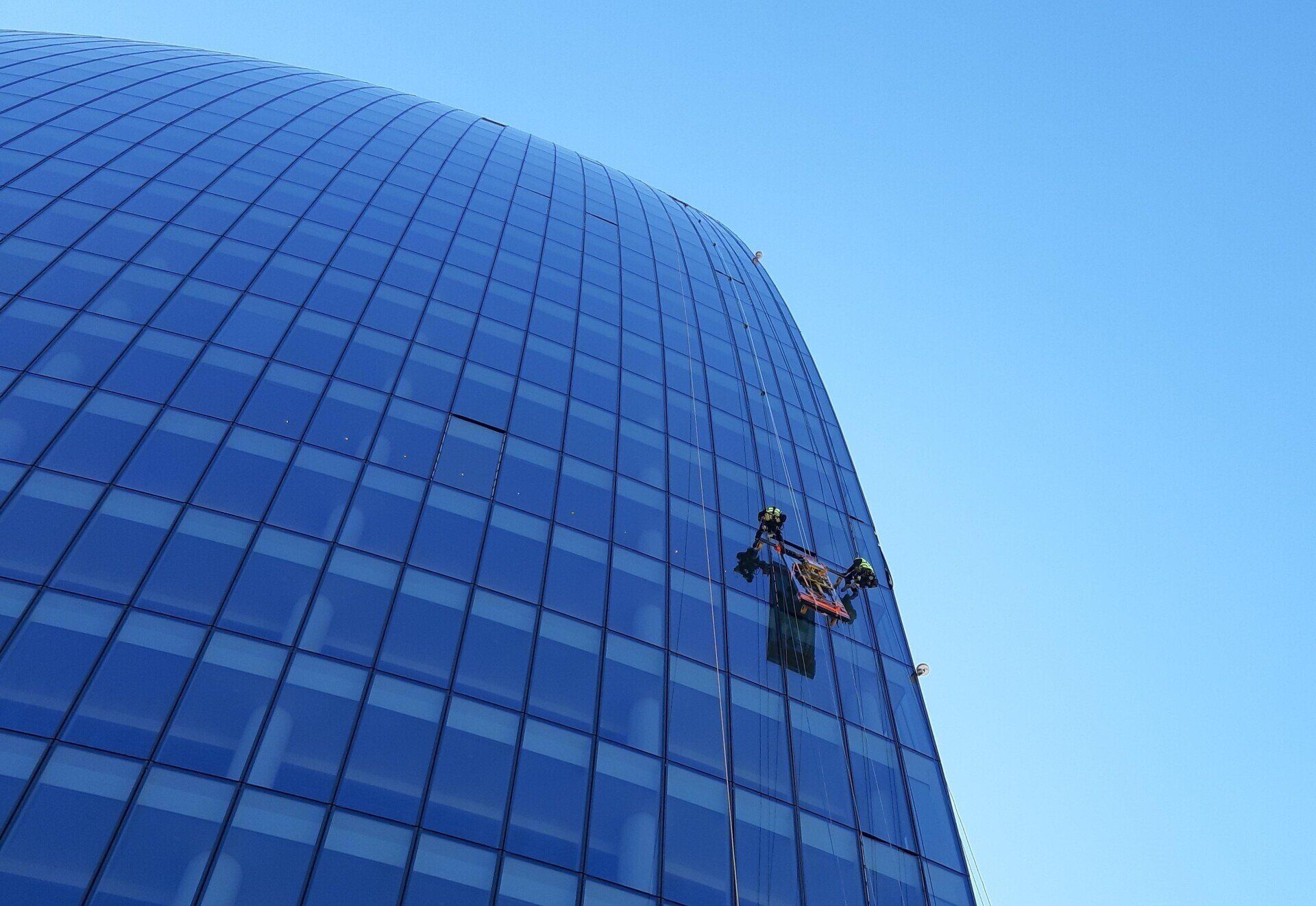 Un lavavetri in ascensore pulisce un alto edificio di vetro blu che si staglia contro un cielo azzurro e terso.