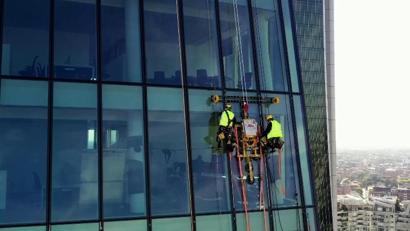 Due lavavetri con i giubbotti gialli sono appesi a una piattaforma sospesa e puliscono la facciata in vetro di un grattacielo.