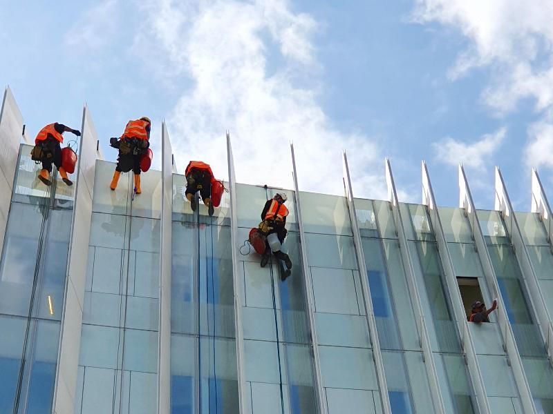 Persone con giubbotti arancioni e caschi che si calano in corda doppia dalla facciata di vetro di un edificio, sotto un cielo nuvoloso.