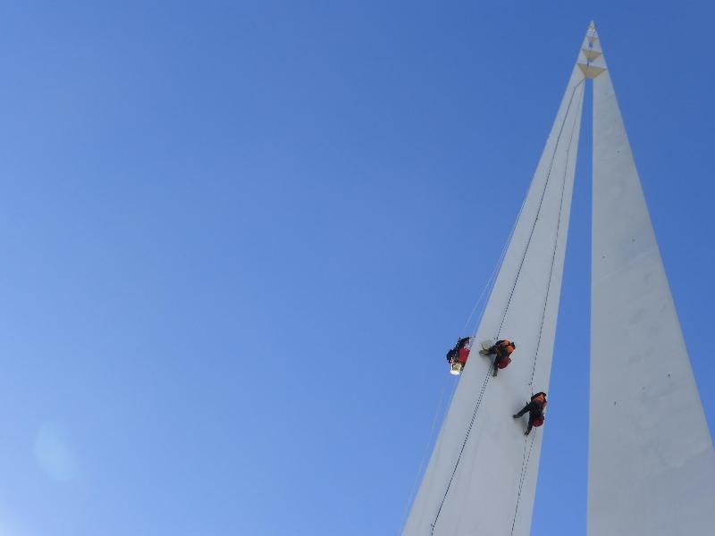 Tre persone scalano un'alta struttura bianca a forma di vela contro un cielo azzurro brillante.