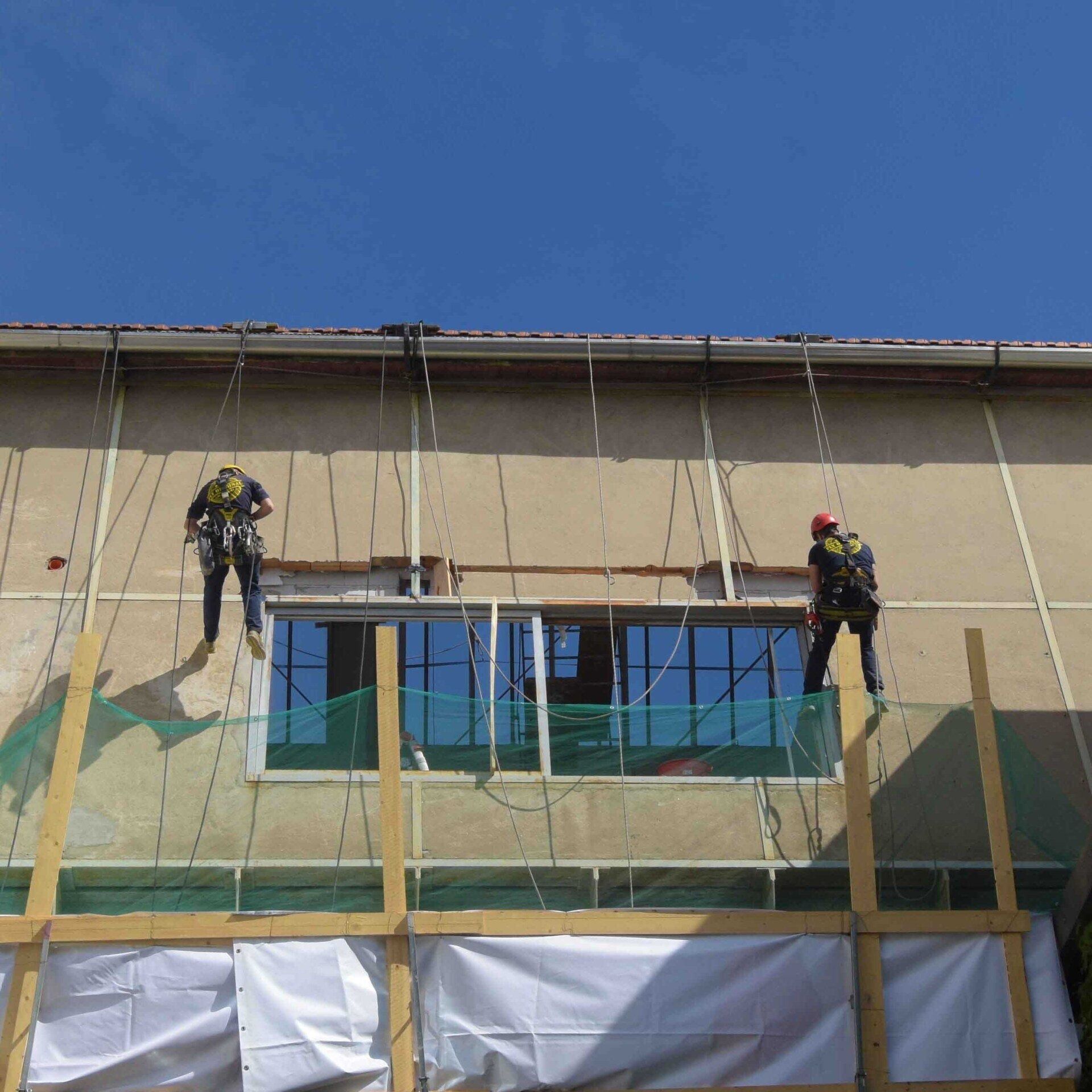Due operai su corde riparano la facciata di un edificio, utilizzando imbracature di sicurezza, con il cielo azzurro sopra.