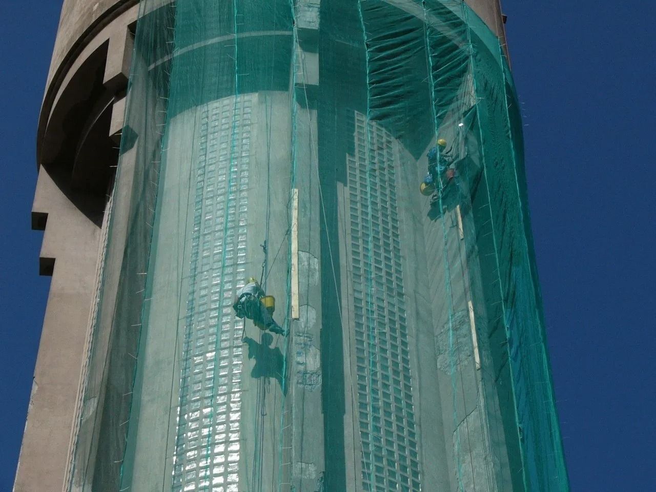 Operai edili su un alto edificio, protetti da una rete verde contro il cielo azzurro.