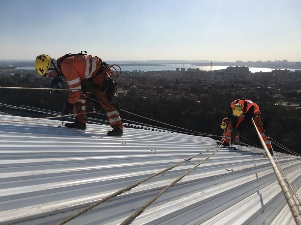 Due persone in tuta arancione su un tetto bianco spiovente, assicurate da corde. La città sullo sfondo.