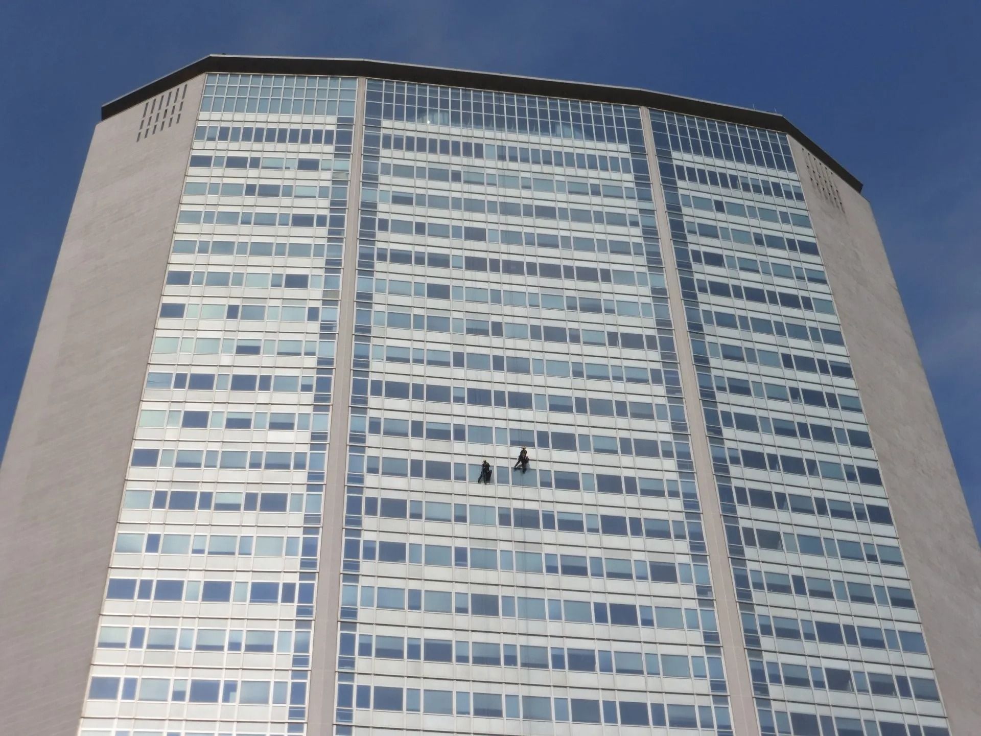 Edificio alto con molte finestre, sono visibili due lavavetri.