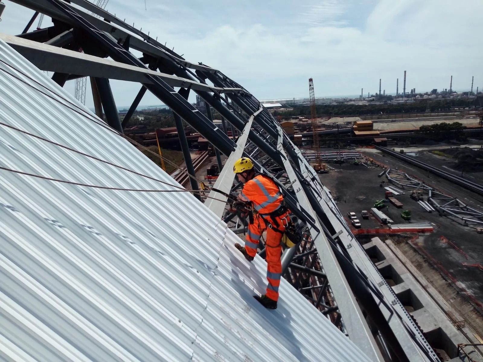 Operaio con equipaggiamento di sicurezza arancione su un tetto metallico inclinato, mentre fissa i cavi. Struttura in acciaio e sfondo industriale.