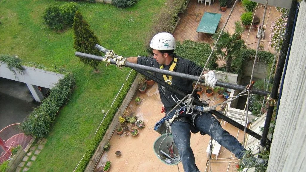 Uomo appeso a un edificio, mentre dipinge. Indossa un casco e un'imbracatura di sicurezza, e tiene in mano un rullo per dipingere. Erba verde visibile sotto.