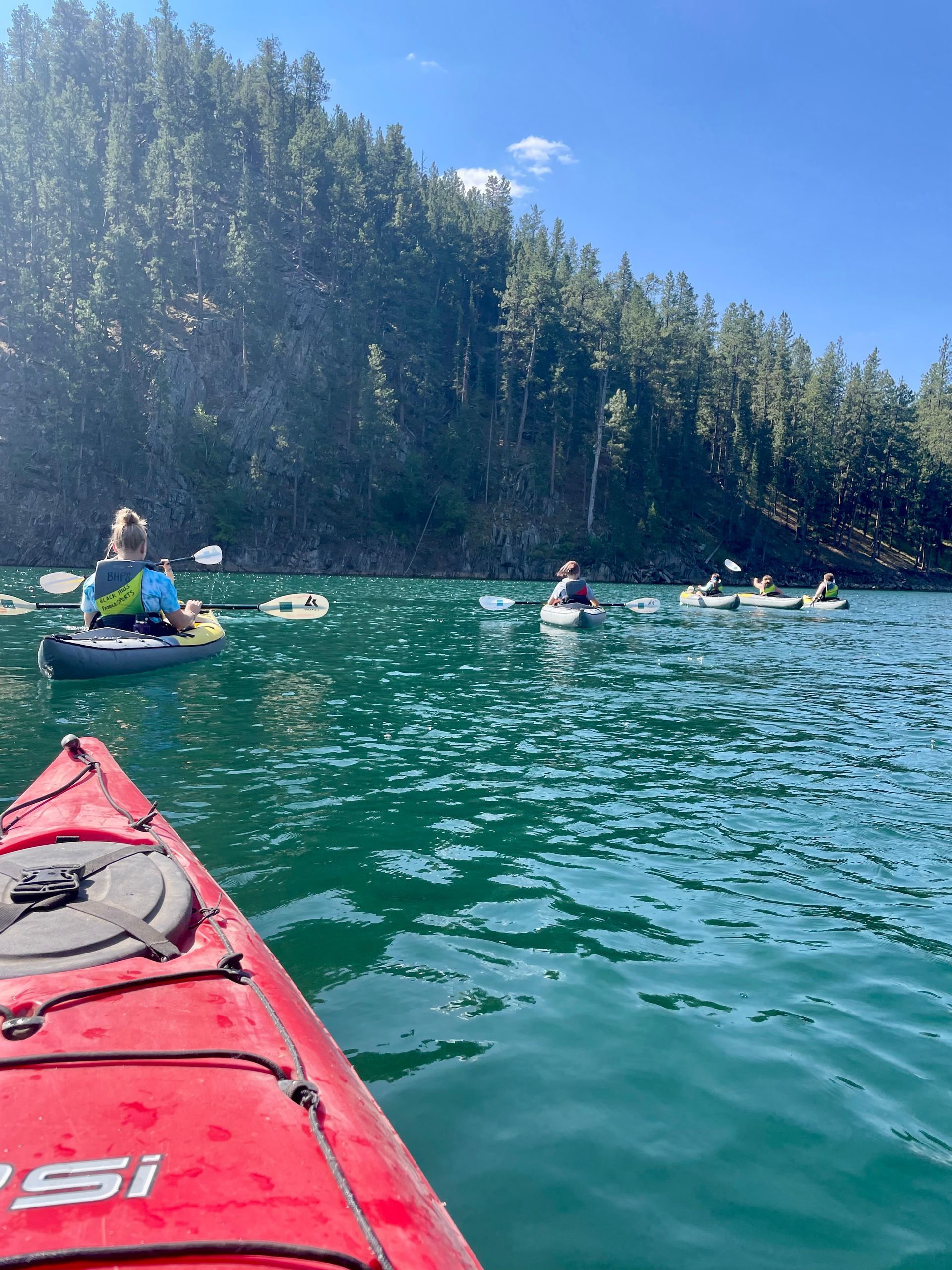 Kayakers on a teal-green lake near a treeline under a blue sky. A red kayak is in the foreground.