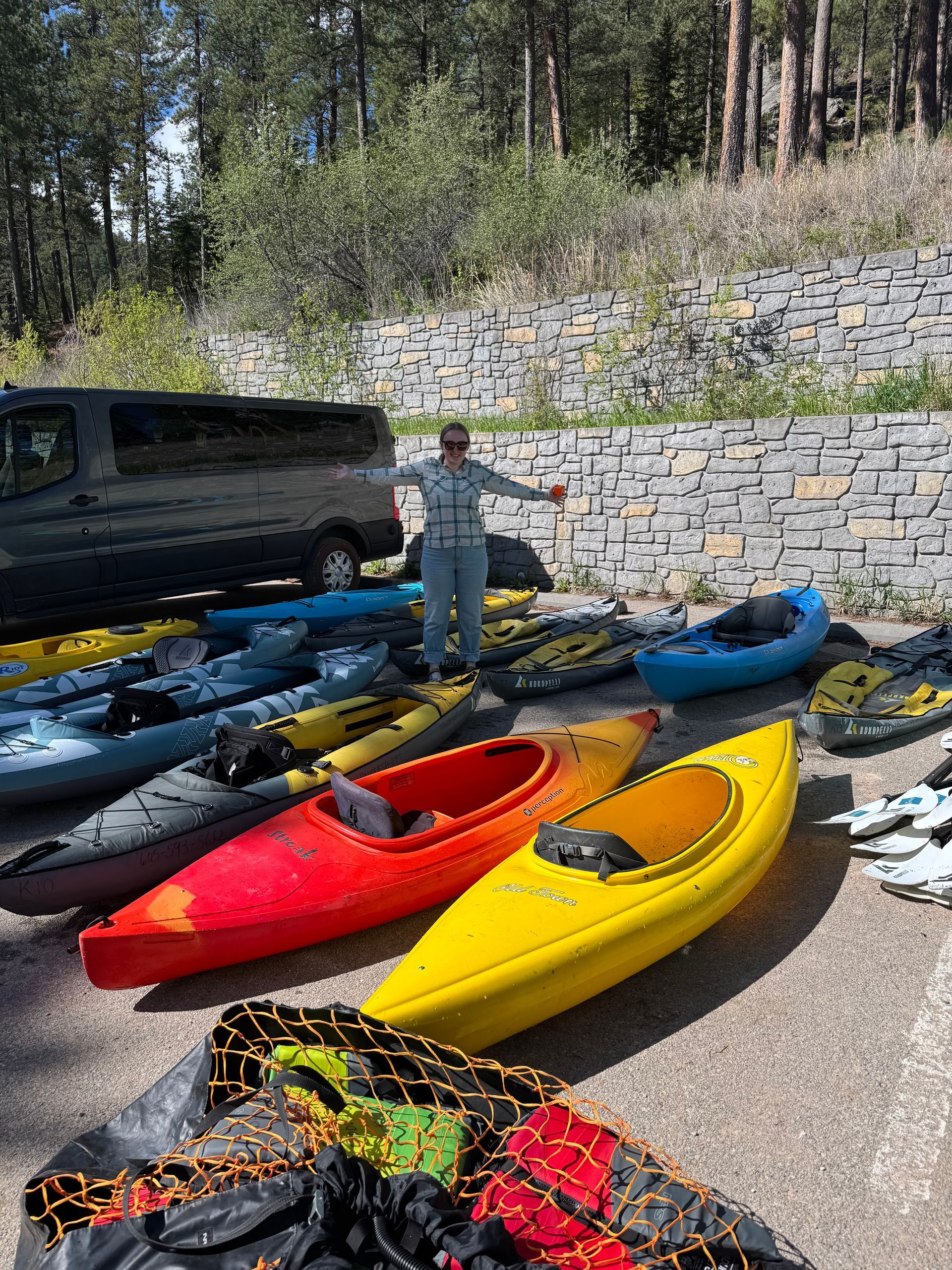 Person stands near many kayaks. Yellow, red, and blue kayaks. Van in background. Outdoor setting.