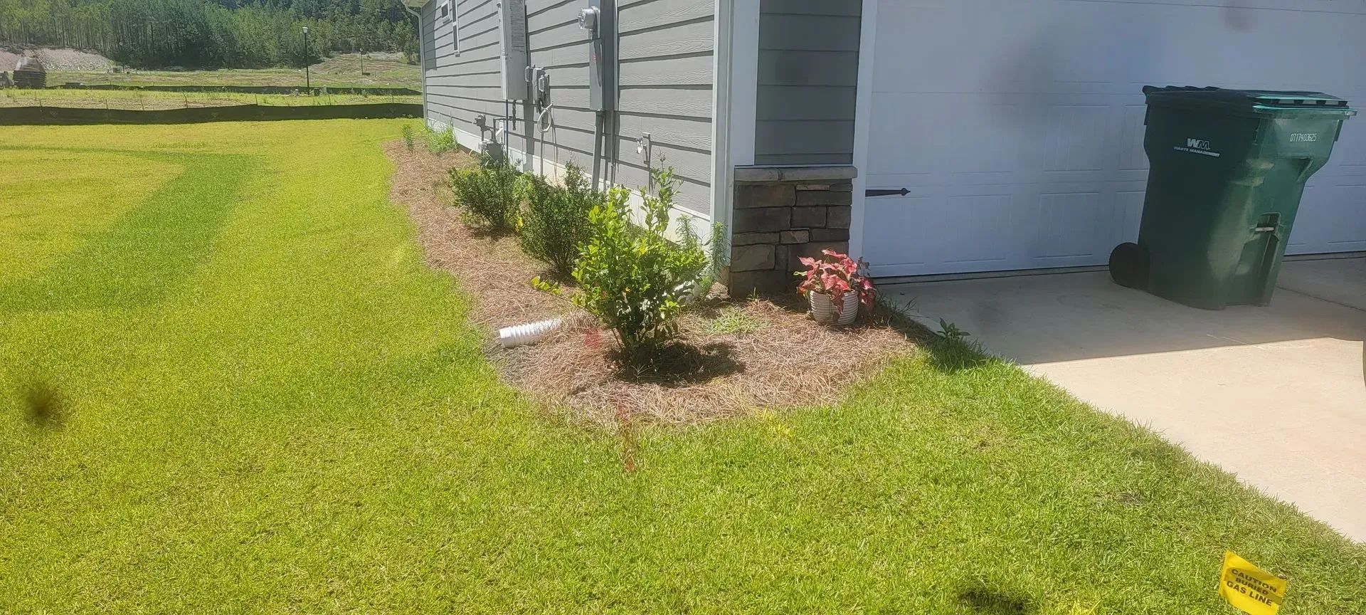 Green lawn beside a house with a flower bed of bushes, a trash can, and a garage door.