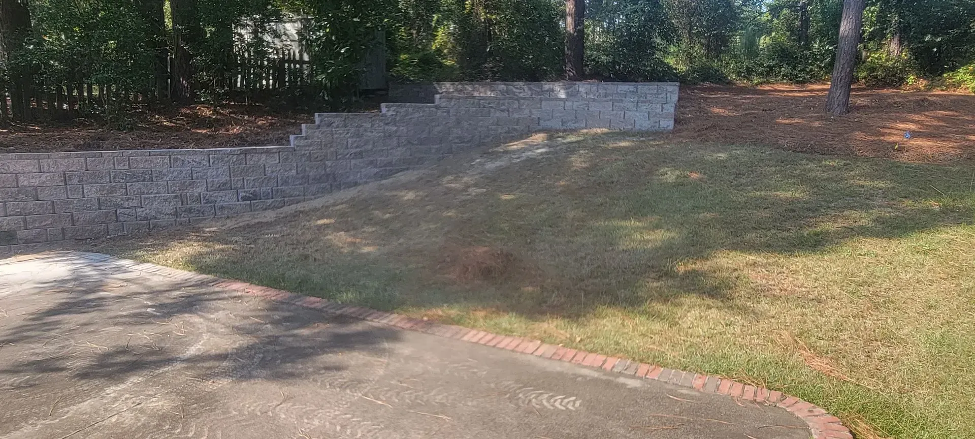 A grassy yard with a retaining wall and trees in the background. A paved area is in the foreground.