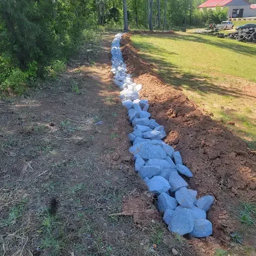 A rock-lined trench for drainage in a grassy yard, with a forest and a house in the background.