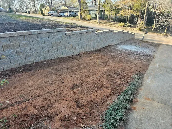 Retaining wall made of gray blocks along a yard with dirt and a paved driveway.