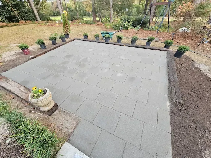 Finished patio of gray pavers surrounded by soil and potted plants; a wheelbarrow sits nearby.